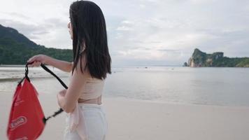 Young asian woman strap on her sling shoulder waterproof bag while walking along beautiful island beach, summer tropical climates travel destination, outdoor adventures gear equipment, island hopping