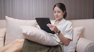 belle femme asiatique confortablement assise sur le canapé à l'aide d'un stylo intelligent touchant une tablette à la recherche d'une boutique en ligne, d'achats de clients en ligne, de confort à la maison, d'achats en ligne de commerce électronique