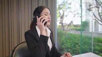 Attractive cheerful sale agency person on contacting customer when sitting at modern glass office. Multi-tasking asian secretary talking on phone arranging confirming appointment, business technology video