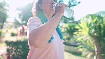 close-up senior aziatische vrouw die water uit een plastic fles drinkt na het sporten in het park, gezondheidszorg voor ouderen, verfrissend drankje, ontspannen pensioen rehydratatie, gezicht afvegen video