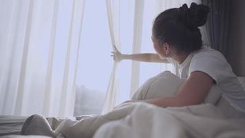 ung vuxen asiatisk kvinna öppet fönstergardin för att se morgonsolljus när man sitter ner i sängen med vackert morgonsolsken, öppna sovrumsgardiner med, utsikt inuti hemboende inhemskt sovrum video