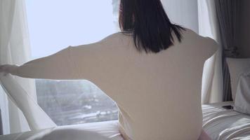 la joven se despierta y se estira en la cama por la mañana, abre las cortinas de la ventana, mira por la ventana con vista al tráfico de la calle de la ciudad, apartamento en el piso alto, nuevo sol que tiene un buen día video