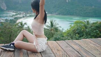 Attractive Asian female tourist sitting down enjoys a natural ocean fresh wind and green tropical green forest on edge of top cliff viewpoint, relaxing weekend active activity, natural eco traveling video