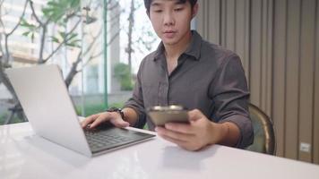Asian freelancer talking to clients and working with the laptop computer at the same time, young adult male using wireless technology for his multitasking works, calling assistant for more information video