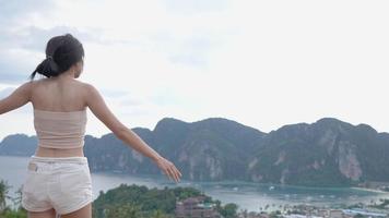 Young woman extended a standing on island viewpoint on top of the hill, beautiful landscape on background. From top view of island mountain cliff, summer vacation travel destination, escape from city video