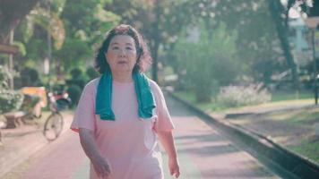 60 years old asian elderly resting after long distances walking in the morning at green fresh park, active grandmother relax walking along concrete paving path, old healthy lifestyle video