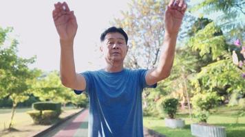 concept de soins de santé senior, homme asiatique d'âge moyen réchauffant son corps en balançant son bras et en marchant avant de courir dans un parc public, journée ensoleillée, entraînement senior, sensation de fraîcheur prévention des maladies âgées video