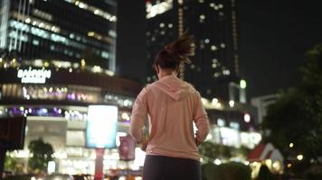 slow-motion, terug volg camera view.female atleet in hooded shirt joggen 's nachts stadsstraten met veel licht op de achtergrond. video