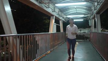 câmera lenta, fonte siga a câmera view.female atleta vestindo um capuz rosa. praticar jogging correndo pelo viaduto à noite todos os dias, as ruas da cidade com muitas luzes. conceito de corrida da cidade à noite. video