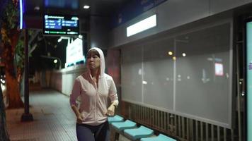 athlète féminine portant un sweat à capuche rose. pratiquez le jogging en courant à travers l'arrêt de bus la nuit tous les jours, les rues de la ville avec beaucoup de lumières en arrière-plan. concept de course de nuit en ville. video