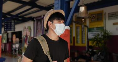 vista lateral del joven viajero asiático caminando y mirando el tren en la estación de tren. hombre con máscaras protectoras, durante la emergencia covid-19. concepto de transporte y viajes.