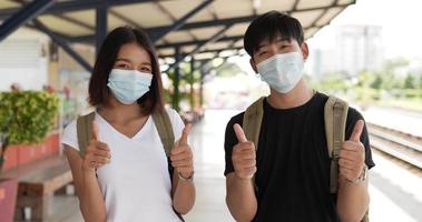 Happy young asian couple show thumbs up look at camera stand in train station. Man and woman wearing protective masks, during Covid-19 emergency. Transportation and travel concept. video