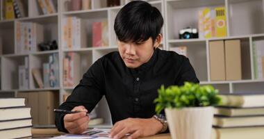 retrato de freelancer focado jovem asiático escrevendo papelada em casa. homem sério trabalhando distante de casa. empresário escrevendo documentos, analisa planos, faz a papelada em um escritório em casa. video