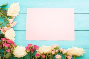 Frame of colorful roses on blue wooden background with paper card and copy space. Top view and selective focus. Mock up photo