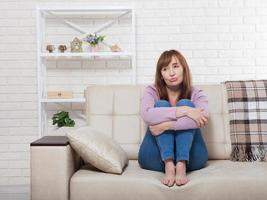 Depressed and sad middle aged woman sitting with clamped knees on bed, coach, sofa at home. Copy space and mock up. photo