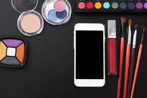 productos cosméticos y accesorios de maquillaje sobre fondo negro. teléfono inteligente con pantalla en blanco. vista superior y espacio de copia. colores de verano foto
