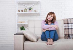 Depressed and sad middle aged woman sitting with clamped knees on bed, coach, sofa at home. Copy space and mock up. photo