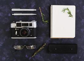 Flat lay, top view office table desk. Desk workspace with retro camera, diary, pen, glasses, case, rosemary on dark background. photo