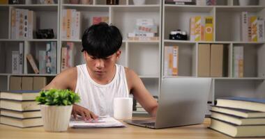 retrato de un hombre asiático cansado en una camiseta usando una computadora portátil y mirando un documento en casa. hombre somnoliento trabajando en casa, pensando y sintiéndose pensativo. trabajar desde casa sobrecargado por la noche en la nueva normalidad. video