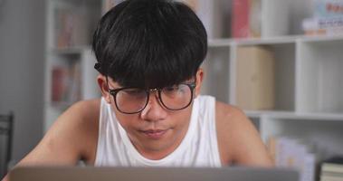 Closeup portrait of Focused freelancer man in singlet and eyeglasses works on laptop at home. Exhausted Asian young man with eyeglasses looks at computer notebook. Work from home and freelance concept video