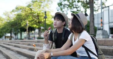 Seitenansicht eines glücklichen asiatischen Paars mit Hut, das Würstchen isst, während es auf der Treppe im Park sitzt. Fröhlicher junger Mann und Frau essen appetitlich. Urlaubs- und Lifestyle-Konzept.