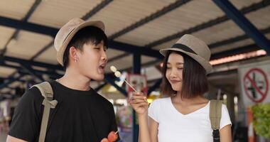 cerca de una pareja de jóvenes viajeros asiáticos comiendo albóndigas en la estación de tren. feliz hombre y mujer hambrientos comiendo aperitivo. concepto de transporte, viajes y comida. video