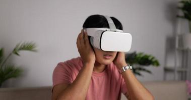 Portrait front view of a Young Asian man sitting on a sofa in the living room wearing a VR headset. Male having VR headset with many emotions, waving their hands in the air. Virtual reality technology video