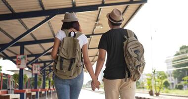 vista posterior de la feliz pareja de viajeros asiáticos con sombrero y mochila mano juntos mientras camina en la estación de tren. sonriente joven y mujer mirando la vista. concepto de vacaciones, viajes y pasatiempos. video