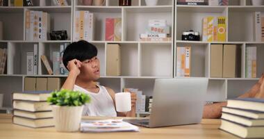 il ritratto di un freelance annoiato in canottiera lavora sul laptop e beve il caffè a casa. il giovane asiatico esamina il taccuino del computer e la gamba sul tavolo. concetto di lavoro da casa. video