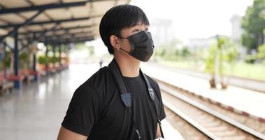 giovane viaggiatore asiatico che scatta una foto sulla fotocamera alla stazione ferroviaria. maschio che indossa maschere protettive, durante l'emergenza covid-19. concetto di trasporto, viaggio e distanziamento sociale.