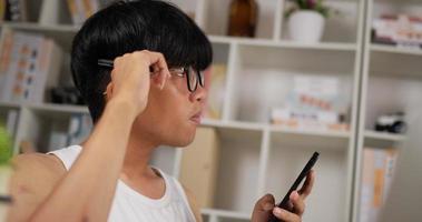 vista lateral de primer plano de los anteojos de un joven asiático usando un teléfono inteligente mientras se sienta en un sofá en casa, un estudiante adolescente usando un teléfono celular moderno navegando por Internet mientras piensa en la habitación. video