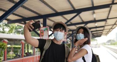 Happy asian traveler couple with hat selfie on smartphone at train station. Young Man taking a pictures on mobile phone. People wearing protective masks, during Covid-19 emergency. Hobby and travel.