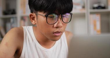 closeup retrato de homem freelancer focado em singlet e óculos funciona no laptop em casa. exausto jovem asiático com óculos Olha para o notebook do computador. trabalhar em casa e conceito freelance video