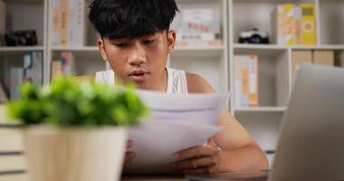 portrait d'un homme asiatique fatigué en singulet utilisant un ordinateur portable et regardant un document à la maison. homme endormi travaillant à la maison, pensant et se sentant pensif. travail à domicile surchargé la nuit dans la nouvelle normalité. video