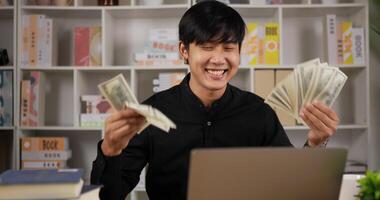 retrato de un hombre asiático feliz que muestra dinero en efectivo a través de una computadora portátil mientras está sentado en el escritorio en la habitación. ganar dinero con internet con negocios en línea, comercio en línea, mercadeo en red, contar dinero. video