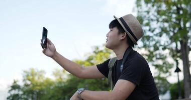 ritratto di vista laterale del giovane viaggiatore asiatico felice con la videochiamata del cappello sul telefono cellulare mentre si siede sulle scale al parco. maschio sorridente che parla sul cellulare al parco. concetto di hobby e stile di vita. video