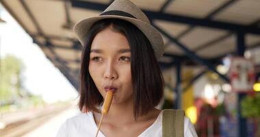 Close up of Young Asian traveler woman with hat eating sausage and looking at camera at train station. Happy hungry female eating appetizer. Transportation, travel and food concept. video