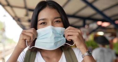 close-up van een jonge aziatische vrouw met kort haar die een beschermend medisch gezichtsmasker draagt en op het treinstation staat. vrouw die beschermende maskers draagt, tijdens covid-19-noodsituatie. reizen en levensstijl. video