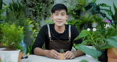 Portrait of a happy young asian male gardener selling online on social media and looking at camera in the garden. Man in face mask video call. Home greenery, selling online and hobby concept
