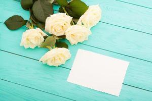 Frame of colorful roses on blue wooden background with paper card and copy space. Top view and selective focus. Mock up photo