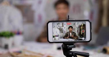Nahaufnahme des Handys einer glücklichen asiatischen Modedesignerin, die eine Skizze von Damenbekleidung zeigt, die Video-Live-Streaming online im Shop aufzeichnet. Kleinunternehmer, Online-Markt-Lieferkonzept. video