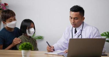 Asian male doctor pediatrician talk to school child girl with face mask make notes in clipboard listen to little patient sit on chair with mother in the hospital. Medical and health care concept.