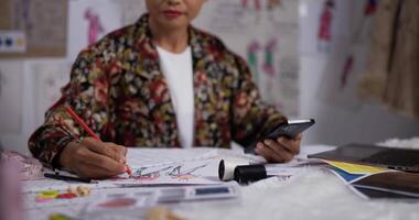 la mano ravvicinata della stilista donna asiatica disegna uno schizzo di abbigliamento femminile mentre guarda lo smartphone in studio. startup imprenditrice sta creando una nuova collezione di vestiti. video