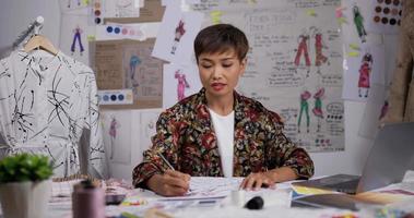 portrait d'une créatrice de mode asiatique dessine un croquis de vêtements pour femmes tout en se tenant au studio. une jeune femme d'affaires réfléchie est en train de créer une nouvelle collection de vêtements. video