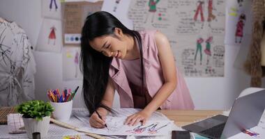 retrato de designer de moda mulher asiática desenha um esboço de roupas femininas em pé no estúdio. feliz empresária de inicialização está em processo de criação de uma nova coleção de roupas. video