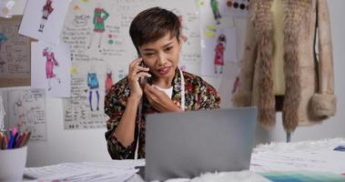 Portrait of Asian woman fashion designer talking on mobile phone with customer while working on laptop in the studio. Happy startup small business owner. SME marketing and entrepreneur concept.