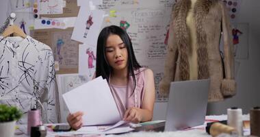 retrato de una diseñadora de moda asiática hablando por teléfono móvil con un cliente mientras trabaja en una laptop en el estudio. propietario de una pequeña empresa de inicio ocupado. pyme marketing y concepto de emprendedor. video