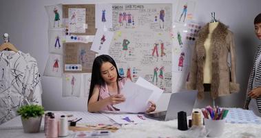 asiatische lehrerin hilft einem modestudentenmädchen, das im studio eine skizze von damenbekleidung sucht. modedesigner, ausbildungs- und bildungskonzept.