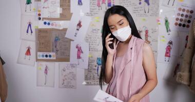 porträt der asiatischen modedesignerin trägt eine maske, die mit dem kunden im studio am handy spricht. designer- und geschäftsinhaberkonzept. video
