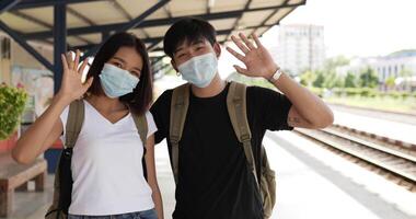 glada unga asiatiska par viftar med handen och tittar på kamerastativ i tågstationen. man och kvinna som bär skyddsmasker under covid-19-nödsituationen. transport och resekoncept. video